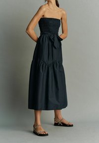 Musta hihaton maksimekko, jossa on smokkauksella koristeltu yläosa, laskostetut helmaosuudet ja rusetti vyötäröllä, yhdistetty hihnallisiin sandaletteihin.