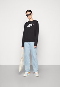 Felpa nera con logo Nike bianco, jeans a gamba larga blu chiaro e sneakers con dettagli arancioni. Tiene una borsa a tracolla beige.