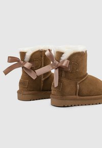 Botas de tobillo de ante marrón con forro de piel sintética blanca y lazos de satén decorativos atados en la parte posterior, exhibidas sobre un fondo gris claro.