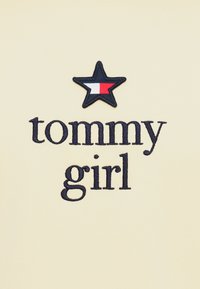 Estrella bordada negra con secciones rojas y blancas sobre texto bordado en negro "tommy girl" en tela beige.