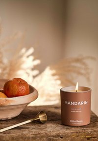 Braune zylindrische Kerze mit der Aufschrift "MANDARIN", angezündet, mit einem goldenen Kerzendocht in der Nähe, neben einer Schüssel mit Orangen auf einer Holzoberfläche.