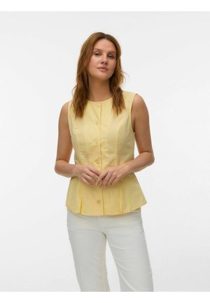 Kobieta o jasnobrązowych włosach, ubrana w żółty bezrękawnik z guzikami i białe spodnie, stojąca na jednolitym białym tle.