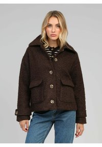 Femme portant un manteau boutonné texturé marron foncé avec des poches à rabat, un large col, sur un jean bleu et un haut rayé.