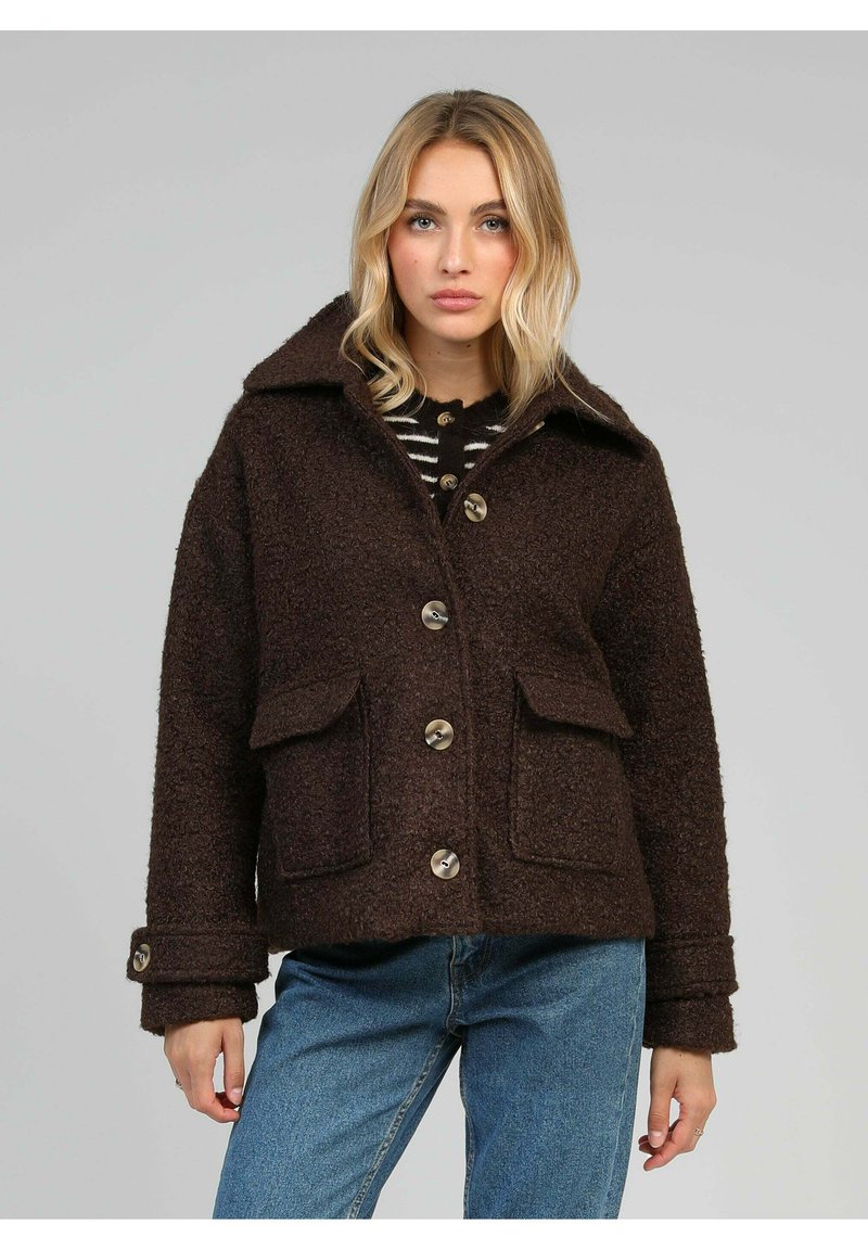 Femme portant un manteau boutonné texturé marron foncé avec des poches à rabat, un large col, sur un jean bleu et un haut rayé.