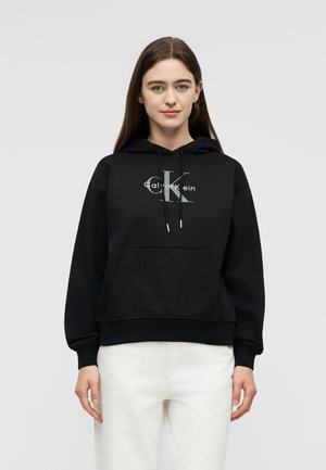 Jeune femme aux longs cheveux bruns portant un sweat à capuche noir Calvin Klein et un pantalon blanc, debout devant un fond gris clair uni.