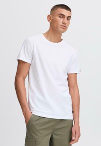 Camiseta blanca de algodón con cuello redondo clásico, mangas cortas y un pequeño logotipo en la manga. Combinada con pantalones cortos de cargador en color verde oliva.
