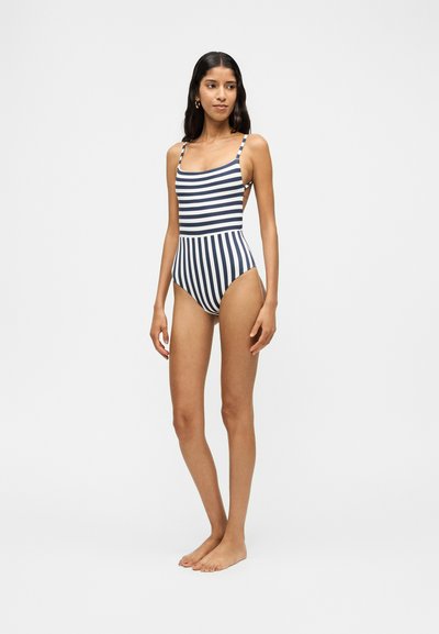 Femme debout pieds nus portant un maillot de bain une pièce avec des rayures horizontales bleu marine et blanches en haut et des rayures verticales en bas.