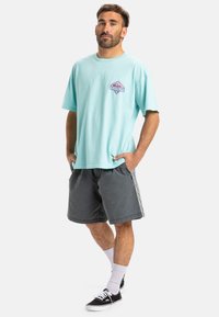 Light blue cotton t-shirt with a purple diamond logo, paired with dark grey shorts featuring a side pattern. Model wears black sneakers and white socks.