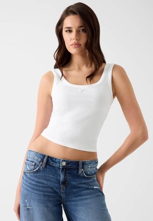 Frau mit langen braunen Haaren, die ein weißes ärmelloses Crop-Top und zerrissene blaue Jeans vor einem schlichten hellen Hintergrund trägt.