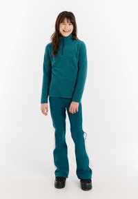 Teal fleece half-zip pullover met lange mouwen, gecombineerd met teal skibroeken met witte zijstrepen. Model draagt zwarte snowboots.