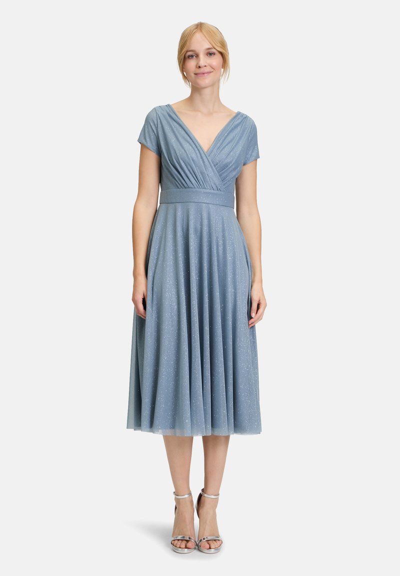 Robe bleu ciel mi-longue avec un décolleté en V, des manches courtes, un corsage plissé et une texture scintillante. Associée à des talons argentés à bride de cheville.