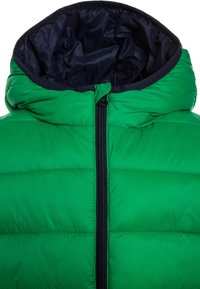 Grön puffjacka med dragkedja fram, huva och marinblå foder. Har quiltade sektioner för isolering och en slät, blank yta.