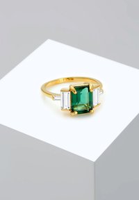 Goldring mit einem smaragdgeschliffenen grünen Stein in der Mitte, flankiert von zwei rechteckigen klaren Steinen, auf einem glatten Band gesetzt.