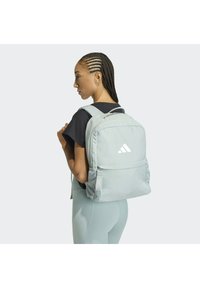 Mochila gris con correas ajustables, cierre de cremallera y un bolsillo frontal. Cuenta con un bolsillo lateral de malla y un logo blanco en la parte posterior.