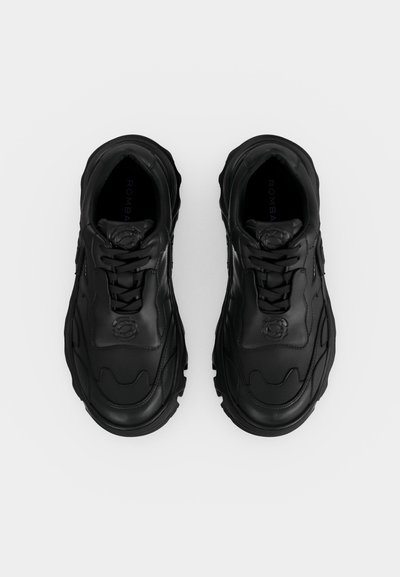 Ζευγάρι μαύρα chunky αθλητικά παπούτσια με κορδόνια, υφασμένο πέλμα και ανάγλυφο λογότυπο στη γλώσσα, σε θέα από ψηλά σε λευκό φόντο.