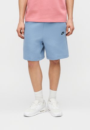 Person iført lyseblå Nike-shorts, pink skjorte, hvide sokker og hvide sneakers, stående på en ensfarvet hvid baggrund.