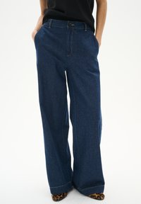 Dunkelblaue, weit geschnittene Denim-Hosen mit Knopfverschluss, Vorderseitentaschen und kontrastierenden Nähdetails. Strukturierter Stoff.