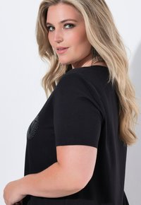 Mujer rubia con ondas sueltas lleva una blusa negra de manga corta con piedras brillantes, pendientes largos de cuentas negras y maquillaje sutil, de perfil.