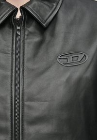 Μαύρο δερμάτινο μπουφάν με έντονο φερμουάρ, μυτερό γιακά και ανάγλυφη λεπτομέρεια λογότυπου στο στήθος. Απαλή υφή με κομψό σχεδιασμό.