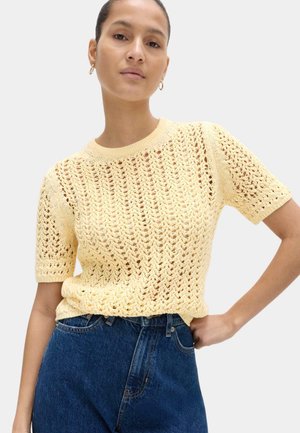 Kobieta ubrana w jasnożółty, krótkorękawowy, szydełkowy top i niebieskie dżinsy z wysokim stanem, stojąca z jedną ręką na biodrze.