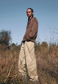 Brun stickad tröja med en avslappnad passform, kombinerad med ljusbeiga baggy byxor. Bakgrunden har högt gräs och sparsamt växtlighet under en blå himmel.