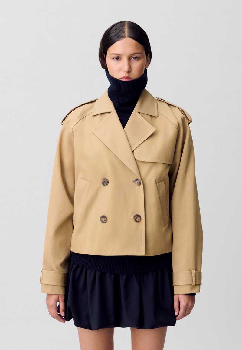 Veste marron à double boutonnage avec de grands boutons, épaulettes et une coupe courte. Portée sur un col roulé noir et une jupe. Texture lisse.