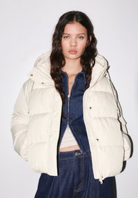 Młoda kobieta o długich ciemnych włosach ma na sobie kremową kurtkę puchową, pod którą nosi ciemnoniebieski jeansowy zip-up i luźne jeansowe spodnie, stojąc na tle jednolitego tła.