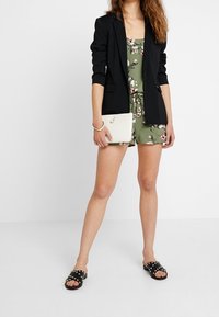 Czarna marynarka na zielonej kwiatowej sukience, trzymająca białą kopertówkę. Czarne klapki z ćwiekowanymi akcentami. Gładkie faktury na całej powierzchni.