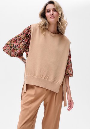 Blonde vrouw in een beige mouwloze gebreide vest over een bloemen top met pofmouwen en beige wijde broek, staand tegen een witte achtergrond.