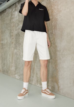 Person iført en sort kortærmet skjorte, hvide knælange shorts, hvide sokker og beige plateau sneakers, som står indendørs.
