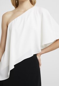 Kobieta w białej bluzie na jedno ramię z asymetrycznym dołem i czarnej spódnicy, trzymająca rękę na biodrze.