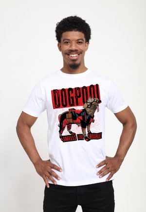 Man smiling with hands on hips wearing white T-shirt showing a cartoon dog in red and black suit with text "DOGPOOL smells like trouble."