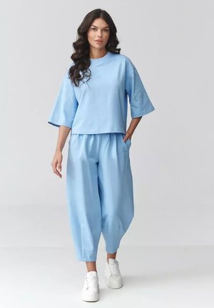 Kobieta o długich ciemnych włosach, ubrana w luźną jasnoniebieską bluzkę i spodnie, idąca do przodu z jedną ręką w kieszeni, białe trampki na jednolitym tle.