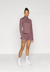 Μπλουζάκι μακρυμάνικο, εφαρμοστό, και σορτς σε μωβ απόχρωση με λευκή επένδυση. Μαλακό ύφασμα, φερμουάρ στον λαιμό, και λογότυπο Nike στο στήθος. Περιλαμβάνονται αθλητικά παπούτσια.