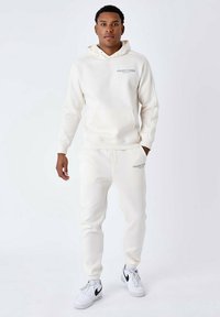 Witte hoodie met een voorzak en logo op de borst, gecombineerd met bijpassende joggingbroek. Beide zijn gemaakt van zachte stof met geribbelde manchetten.