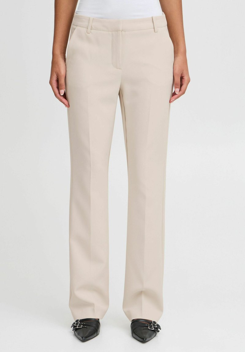 Femme portant un pantalon beige à jambes droites et des chaussures noires à bout pointu avec des brides à double boucle, se tenant devant un fond uni.