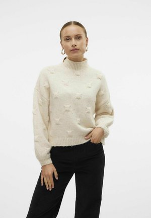 Pull crème avec un col montant, motif texturé et coupe ample, associé à un pantalon en velours côtelé noir. Détail en tissu tricoté doux.