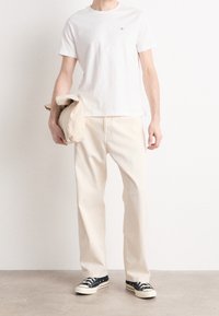 Bílá bavlněná tričko, béžové bavlněné kalhoty, drží složenou béžovou věc. Černobílé tenisky. Jednoduchý, ležérní outfit se neutrálními barvami.