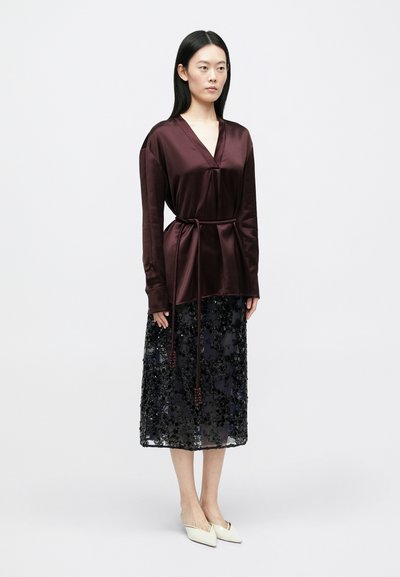 Γυναίκα που φοράει σκούρα βουργουνδί σατινέ μπλούζα με ζώνη και μαύρη μίντι φούστα με λουλουδάτα παγέτα, στέκεται μπροστά σε απλό λευκό φόντο.