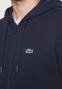 Sudadera con cremallera de color azul marino, hecha de una tela suave, que presenta un logotipo bordado de cocodrilo verde en el lado izquierdo del pecho y cordones ajustables.