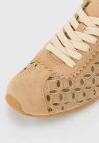 Barna velúr sneaker krémszínű fűzővel, bonyolult kivágott mintázattal és texturált részletekkel. Lekerekített orral és gumitalppal.
