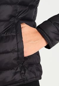 Détail d'un manteau matelassé noir montrant une main dans une poche. Caractéristiques : design matelassé, texture lisse et poignets ajustés.