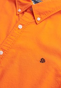 Pomarańczowa koszula z kołnierzykiem, wykonana z bawełny, z zapięciem na guziki i niebieskim wyszywanym logo na piersi. Gładka tekstura.