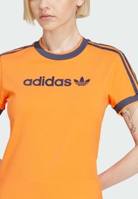 Oranžs kokvilnas T-krekls ar apaļu kakla izgriezumu, pelēki akcenti uz piedurknēm un zilā Adidas logo ar trefoil virs teksta.