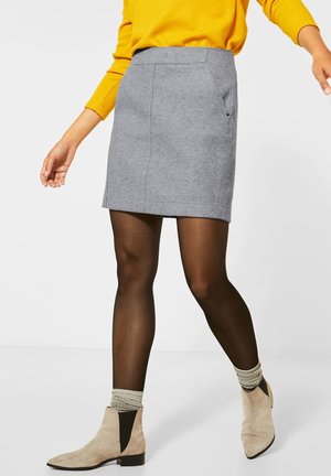 Jupe grise en tissu texturé, avec une taille ajustée et des poches sur les côtés, portée avec des collants noirs et des bottines beige avec un panneau contrastant.