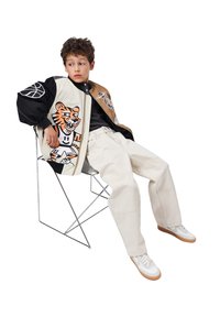 En ung dreng sidder på en metalstol iført en multicolor jakke med tiger- og basketball-designs, beige bukser og hvide sneakers, og kigger sidelæns.