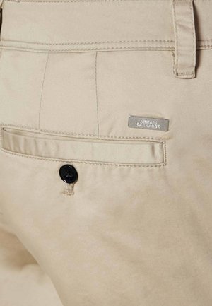 Poche de pantalon beige avec bouton noir et étiquette en métal du logo Armani Exchange au-dessus de la poche du côté droit.