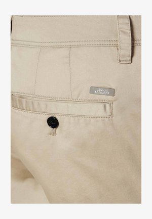 Poche de pantalon beige avec bouton noir et étiquette en métal du logo Armani Exchange au-dessus de la poche du côté droit.