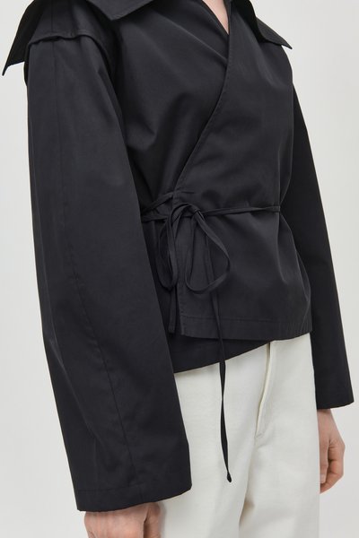 Veste noire style cache-cœur avec large col et manches longues, nouée à la taille avec de fines lanières noires, associée à un pantalon blanc.