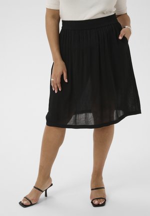 Frau trägt einen schwarzen knielangen Rock mit elastischem Bund und schwarze Sandalen mit Absatz, Hand in der Tasche ruhend.